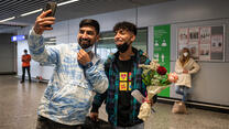 In an airport, two young men look at a phone to take a selfie. They are smiling and one of them holds flowers. 