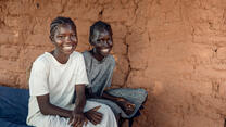 Two young girls sit on the edge of a bed and smile looking at the camera. 