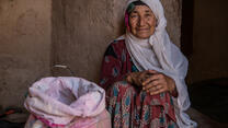 A smiling elderly Afghan woman sits on the ground with her hands folded at her knees with a large sack grain beside her.