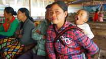 A mother in Myanmar sits holding her yawning baby in a sling. 