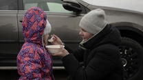 Dressed in winter clothes, a mother kneels down outside to feed her young daughter. 