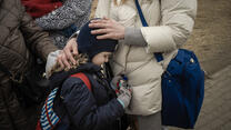 A close up of a young girl leaning against her mother, who places her hands on her back and head. 