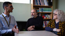 Shir Zad Sarbaz gestures as he speaks while sitting at a table with Mandy Patinkin and Kathryn Grody, who are listening. 