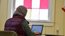 A young woman, with her back to the camera, sits at a desk and types on her laptop.