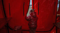 A young girl wearing a red winter coat stands in a red tent, peering out.
