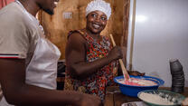 Julia, 42, a baker from South Sudan smiles as she stirs ingredients in a bowl looking at her mentor Joshua who stands smiling beside her.