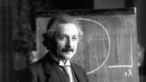 Albert Einstein stands near a blackboard, smiling