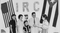 A Cuban family of four is greeted by an American IRC worker, standing in front of a wall over which a Cuban flag and a U.S. flag hang side-by-side