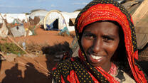 A smiling young woman stands outside a tent in Dadaab refugee camp in Kenya on a sunny day,