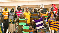 A group of women who are part of an IRC livelihoods program in Nigeria stand outside, lifting hand-knitted tops to show them off.  