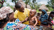 An IRC health worker holds a crying baby, giving him a medical checkup in a village in Central African Republic as his mother looks on smiling. 