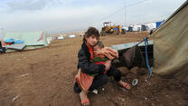 A displaced girl sits just outside a tent in Dohuk, Iraq holding a young child in her arms, bundled in a blanket.