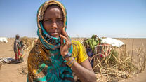 Deqa Omer, 32, stands among makeshift tents in a parched landscape in Ethiopia during a drought, her fingers resting on her cheek as she looks at the camera.