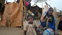 Sudanese refugee woman and girl sitting outside a shelter made of blankets and sheets.