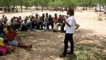 A woman who is an IRC staff member stands speaking animatedly to a group of people seated on the ground in the shade of a tree in Zimbabwe