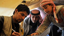 A volunteer from the International Rescue Committee holds a pen while two Syrian refugees point to an item on the page the volunteer is writing on.