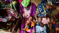 Malian women wearing brightly colored clothing hold children as they are measured and weighed.
