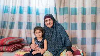 A young girl and her grandmother sit at the edge of a bed and pose for a photo.