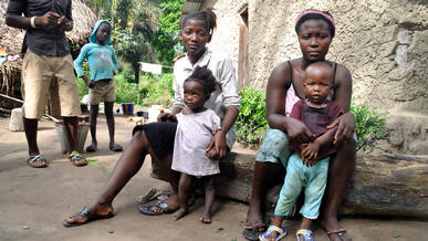 Two women sit on a bench and hold their young children, while a man and a girl stand in the background.