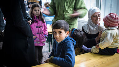 A young boy sits at a table in a refugee shelter in Serbia, surrounded by other children and their families seeking safety in Europe.