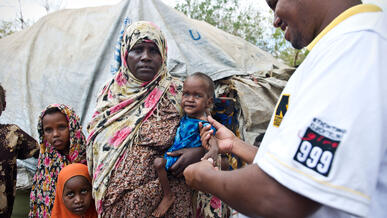 A male IRC health worker in Dadaab refugee camp stands outside a tent examining a baby girl being held in her mother's arms for signs of malnutrition.