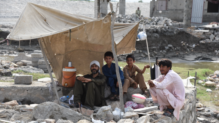 A man and 3 boys sit outside in a makeshift tent. They sit on a pile of rubble and in the background is their destroyed home.