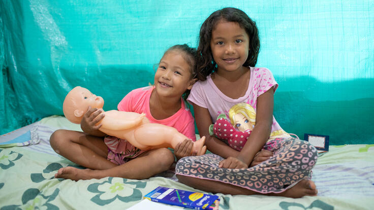 Two girls sit together smiling and holding dolls.