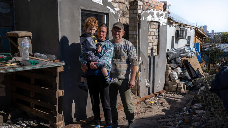 Marina stands next to her husband while holding their son. In the background is their home that was destroyed by the war.