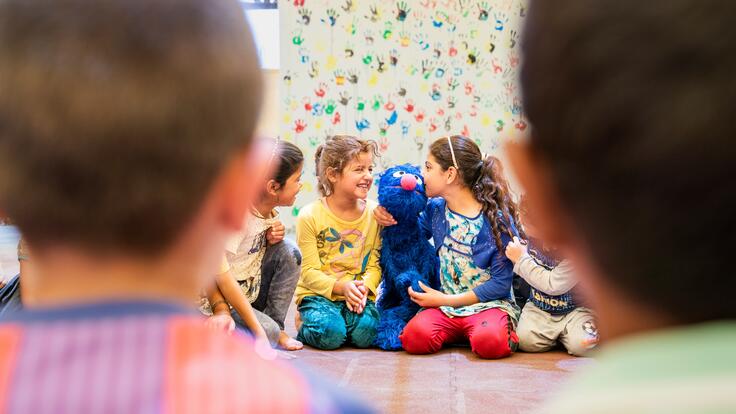 Refugee children sit on the ground in a circle playing witha puppet of the Sesame Street character Grover, a blue monster.