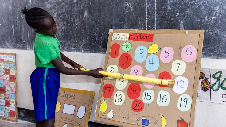 A young student stands at the front of a classroom, pointing at numbers on a board.