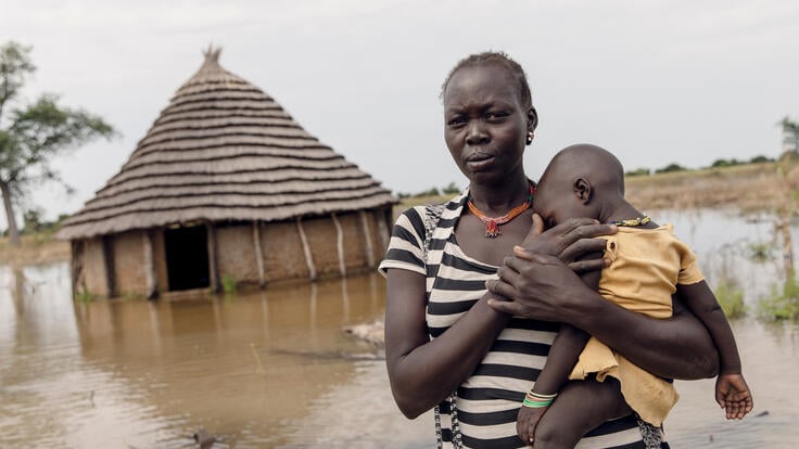 Översvämningar i Sydsudan - kvinna håller i sitt barn framför sitt översvämmade hus. 