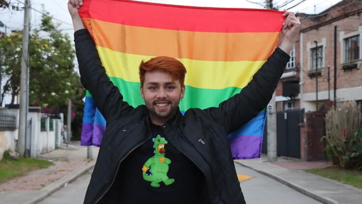 Jesús Ruiz lächelt in die Kamera und hält hinter sich eine Pride-Fahne hoch