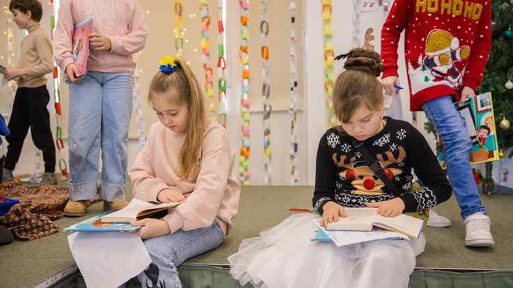 Two girls sit side by side, reading new books. Behind them, others play during an IRC book distribution event.