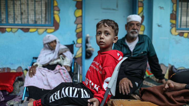 A Palestinian boy sits in a chair at a UNRWA school. An elderly woman and man sit nearby.