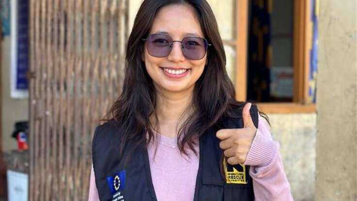 A young woman with long brown hair and sunglasses is looking at the camera and doing a thumbs up with her left hand