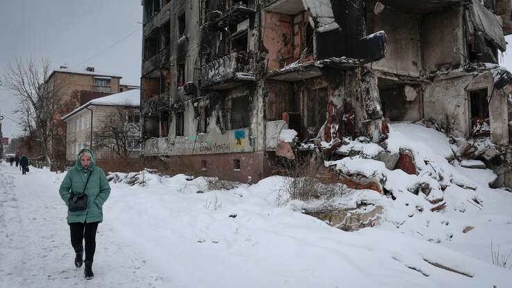 A woman walks down a snow-covered street in Ukraine, beside a building that has been destroyed in the war/