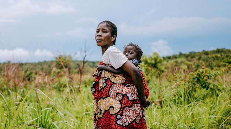 En mamma som bär sitt barn på ryggen ståendes på ett fält. 