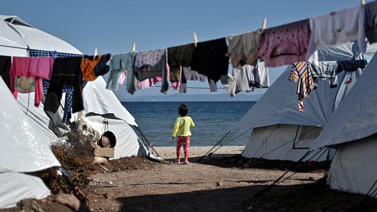 A young child, standing between tents and underneath a line of washing, looking out at the sea