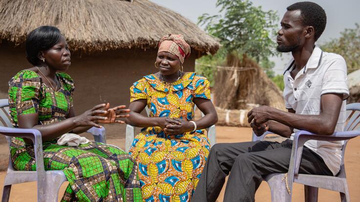 Two members of a women's group sit on plastic chairs outside a house in a Uganda refugee settlement speaking with a man about women's rights