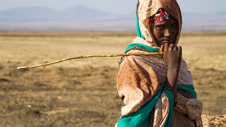 A teenage girl, holding a stick and looking at the camera, stands in a drought affected very dry landscape with mountains in the distance. 