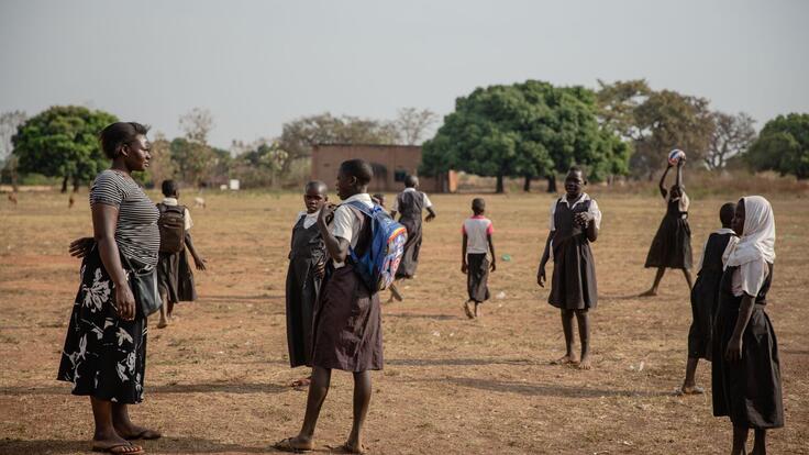 Jackie stands in a field with young girls in school uniforms. One holds a ball as they prepare to play netball. 