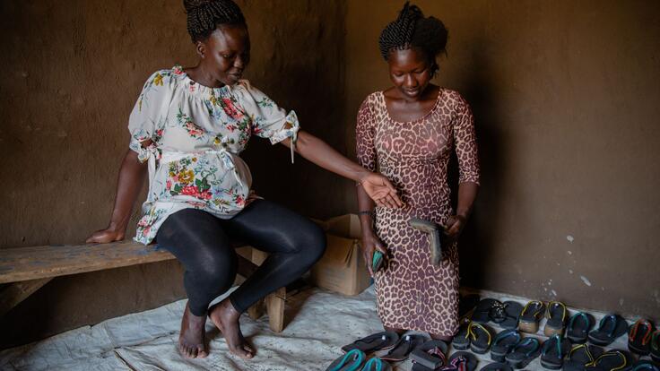 Grace sits on a bench next to a refugee named Vivian, who is kneeling next to shoes arranged neatly on the ground. She is holding one of the shoes and looking down at them.  