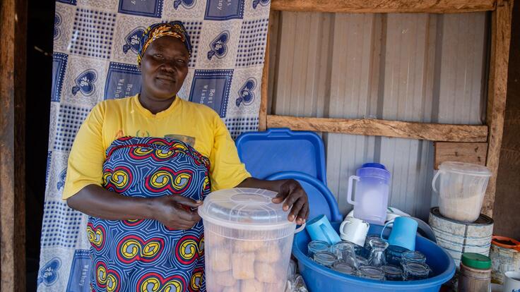 Jemimah poses with the cakes she sells to her community. She is wearing an apron and holding a box of the cakes, looking at the camera. 
