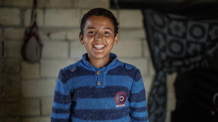 Tareq, wearing a blue and white sweater, smiles for the camera in his home. 