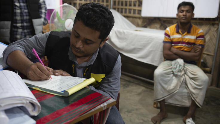 Dr. Mahmudul Hossain, wearing an IRC vest, sits at a desk writing notes. A patient sits behind him. 