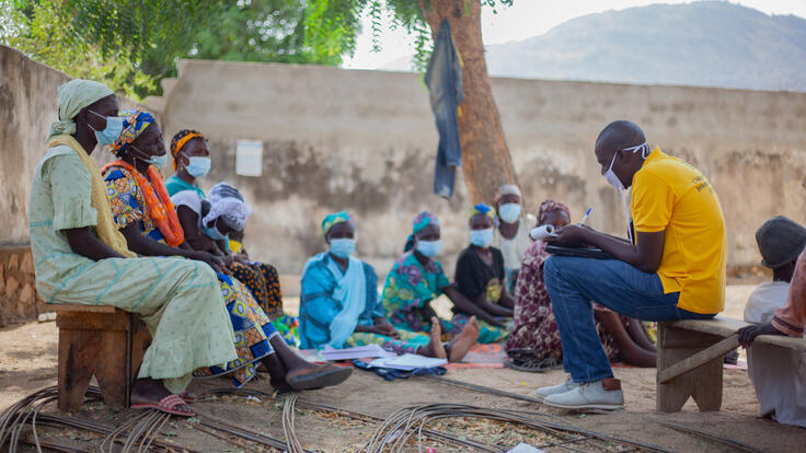 54-year-old Zara Tapita sits on a wooden bench with other parents listening to an IRC staff member leading a session for caregivers, to help them ensure their children can learn and thrive despite crises.