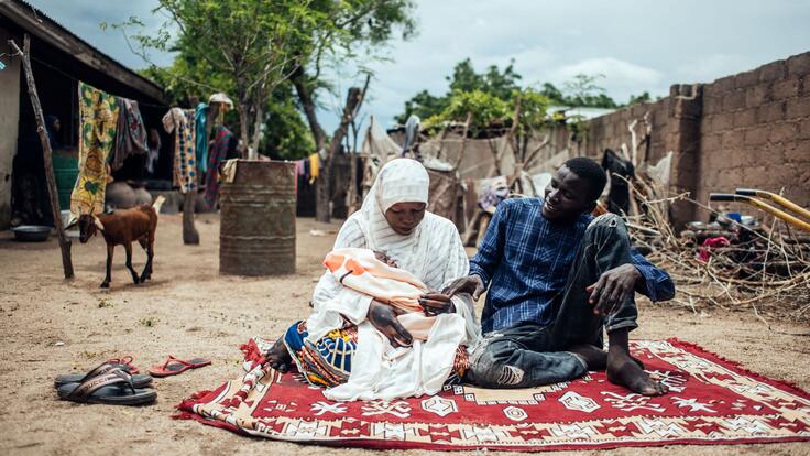 A Nigerian man and woman sit on a rug outside holding their baby.