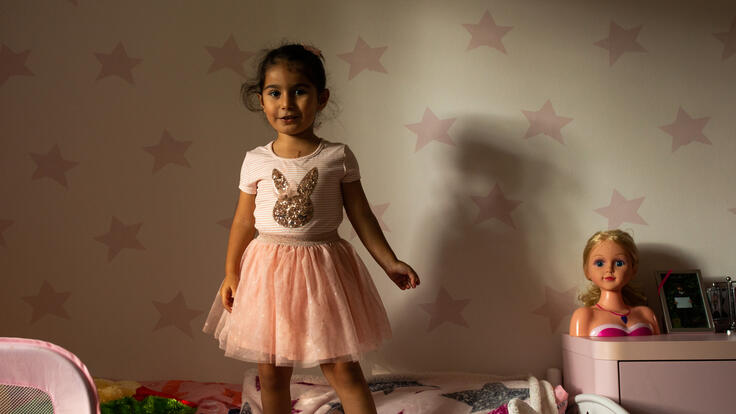 Four-year-old Nasrin stands smiling on her bed with toys around her.