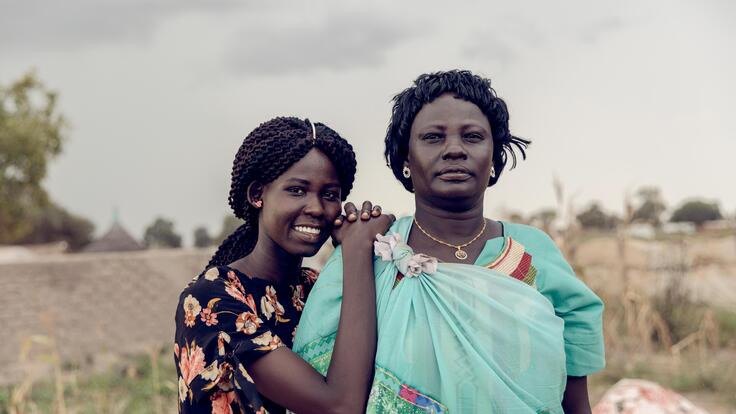 A young girl poses with her hands on one of an older womans shoulders, smiling. Both look straight at the camera.