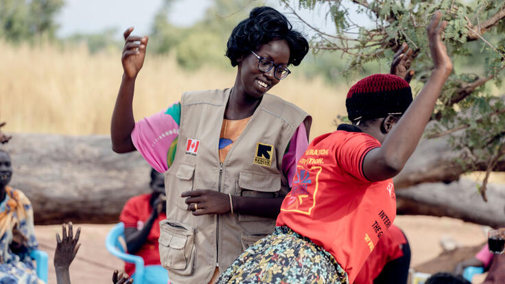 Two women, one wearing an IRC-branded vest, link arms and dance. 